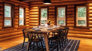 A wooden log cabin dining area with a table set for six, surrounded by chairs, and large windows with a view of trees. - Perry River Lodge, Your Riverside Retreat (Columbia-Shuswap)