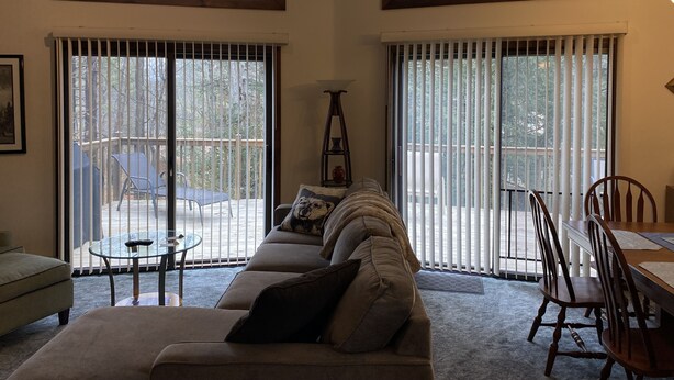 A living room with a sofa, a small table, and a dining table with chairs. There are large windows with blinds, and a balcony is visible through the windows. - Cozy Lakefront Home; Close to skiing and waterparks (Blakeslee)