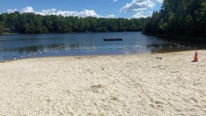 A sandy beach with a dock extending into a calm lake, surrounded by dense forest. - Cozy Lakefront Home; Close to skiing and waterparks (Blakeslee)