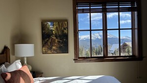 Une chambre d’hôtel avec un lit, un tableau représentant un sentier de forêt, une fenêtre donnant sur une vue de montagne et une lampe.