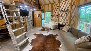 A cozy interior with a wooden ladder, a sofa, a large rug, and a dog. - Windsong yurt, in the treetops, hottub on the top deck, petfriendly, stargazing (Hillsville)