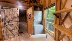 A compact room with a bed, a washing machine, and a window with a view of greenery. - Windsong yurt, in the treetops, hottub on the top deck, petfriendly, stargazing (Hillsville)
