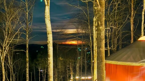 A treehouse-style cabin with a curved roof, surrounded by tall trees and illuminated by string lights. - Windsong yurt, in the treetops, hottub on the top deck, petfriendly, stargazing (Hillsville)