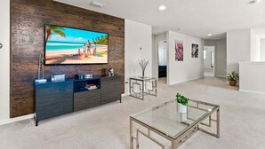 A modern living room with a flat-screen TV mounted on a wooden wall, a glass coffee table, and a minimalist dining area. - Family Paradise Close to Disney w Resort Amenities (Kissimmee)