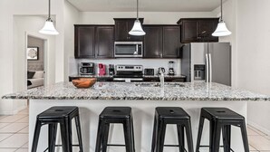 A modern kitchen with dark cabinets, a granite countertop, a central island with stools, and overhead pendant lighting. - Family Paradise Close to Disney w Resort Amenities (Kissimmee)
