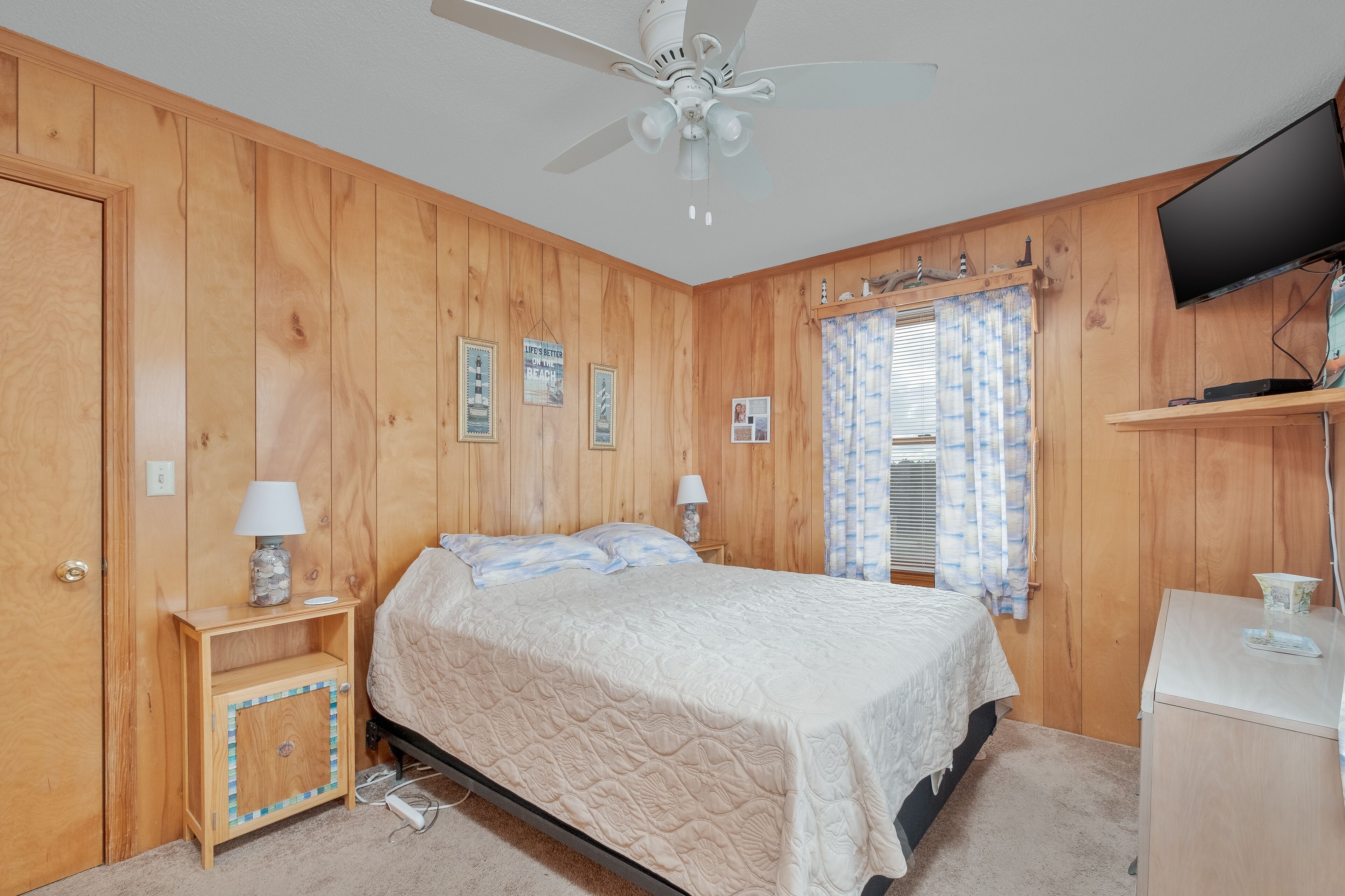 A bedroom with wooden paneling, a bed with a quilt, two bedside lamps, a ceiling fan, a television on a stand, and a window with curtains.