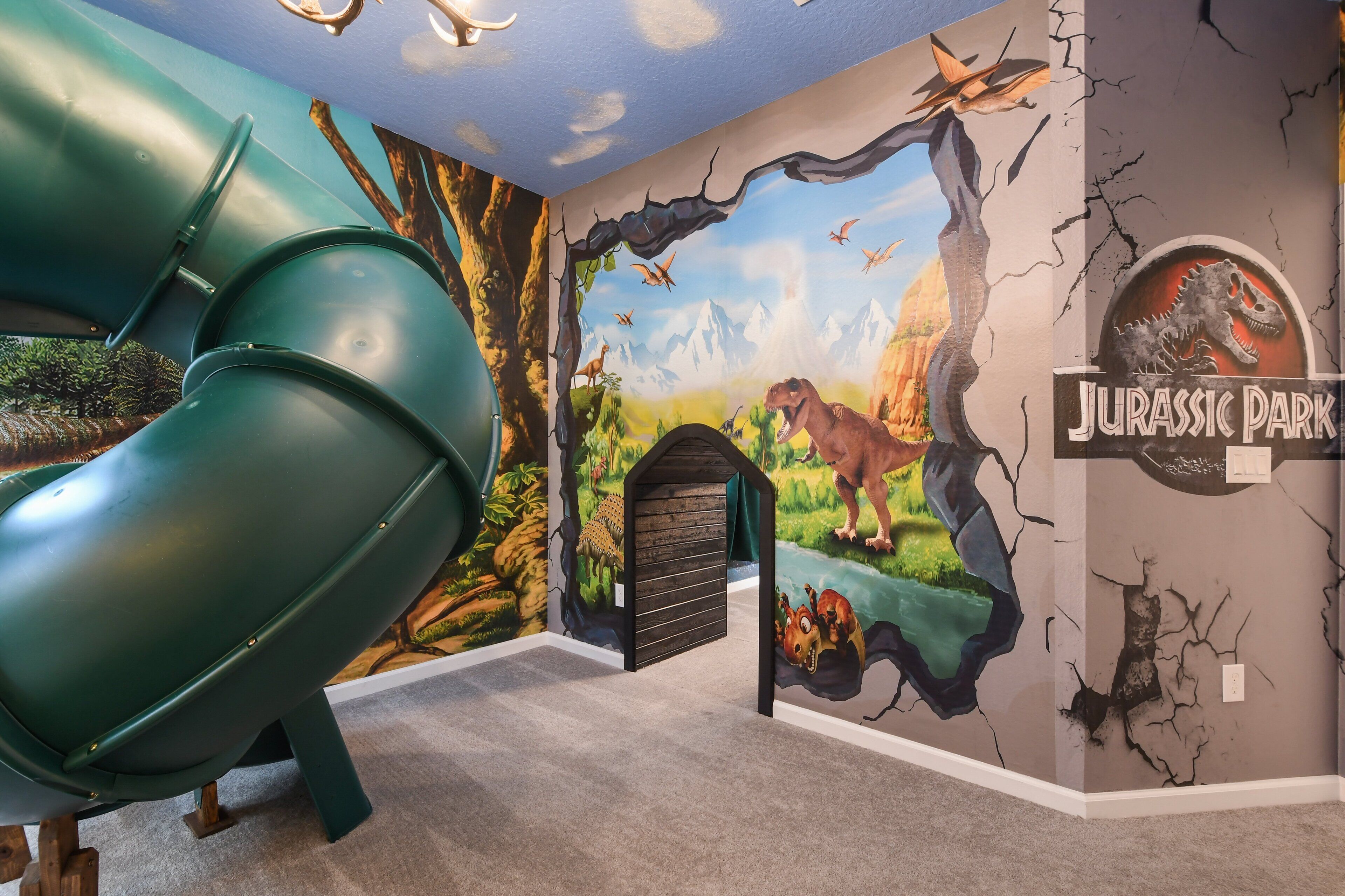 A room with a green slide, a mural of dinosaurs, and a 'Jurassic Park' sign.