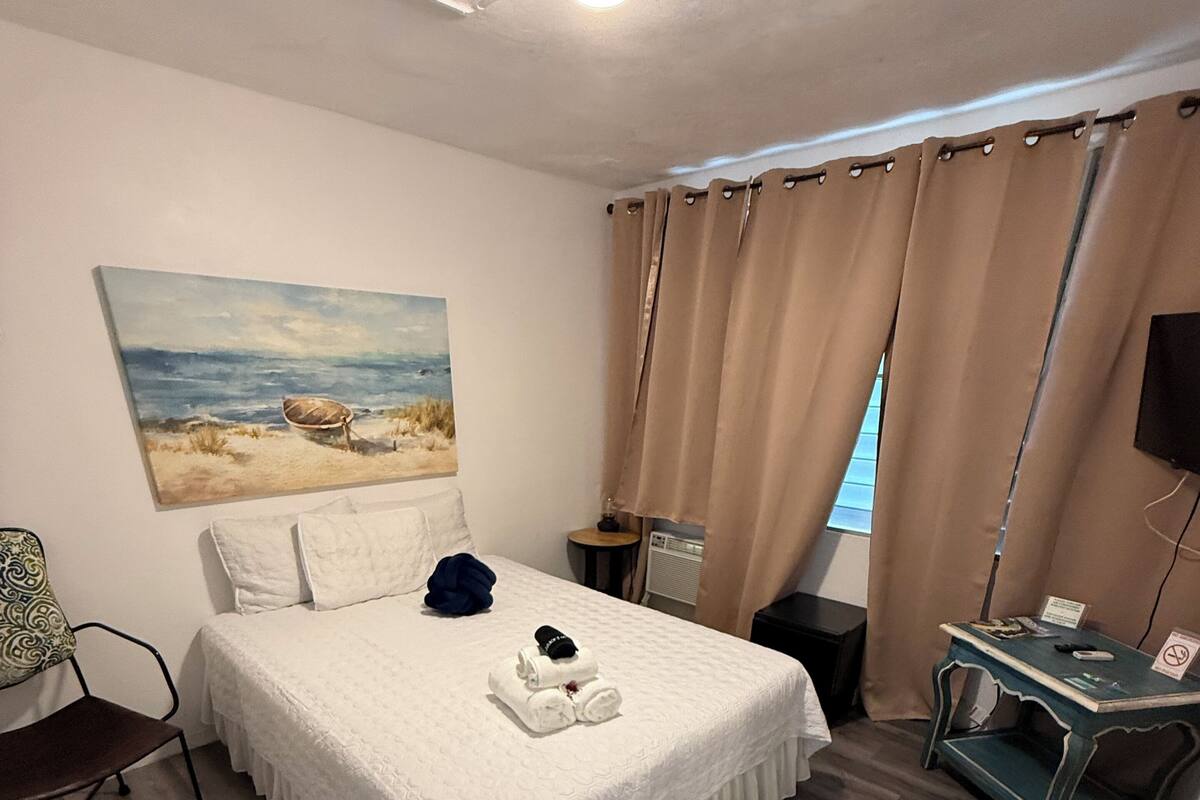 Ein ordentlich eingerichtetes Schlafzimmer mit Bett, Schreibtisch, Stuhl, einem Bild an der Wand und einem Fenster mit Vorhängen.