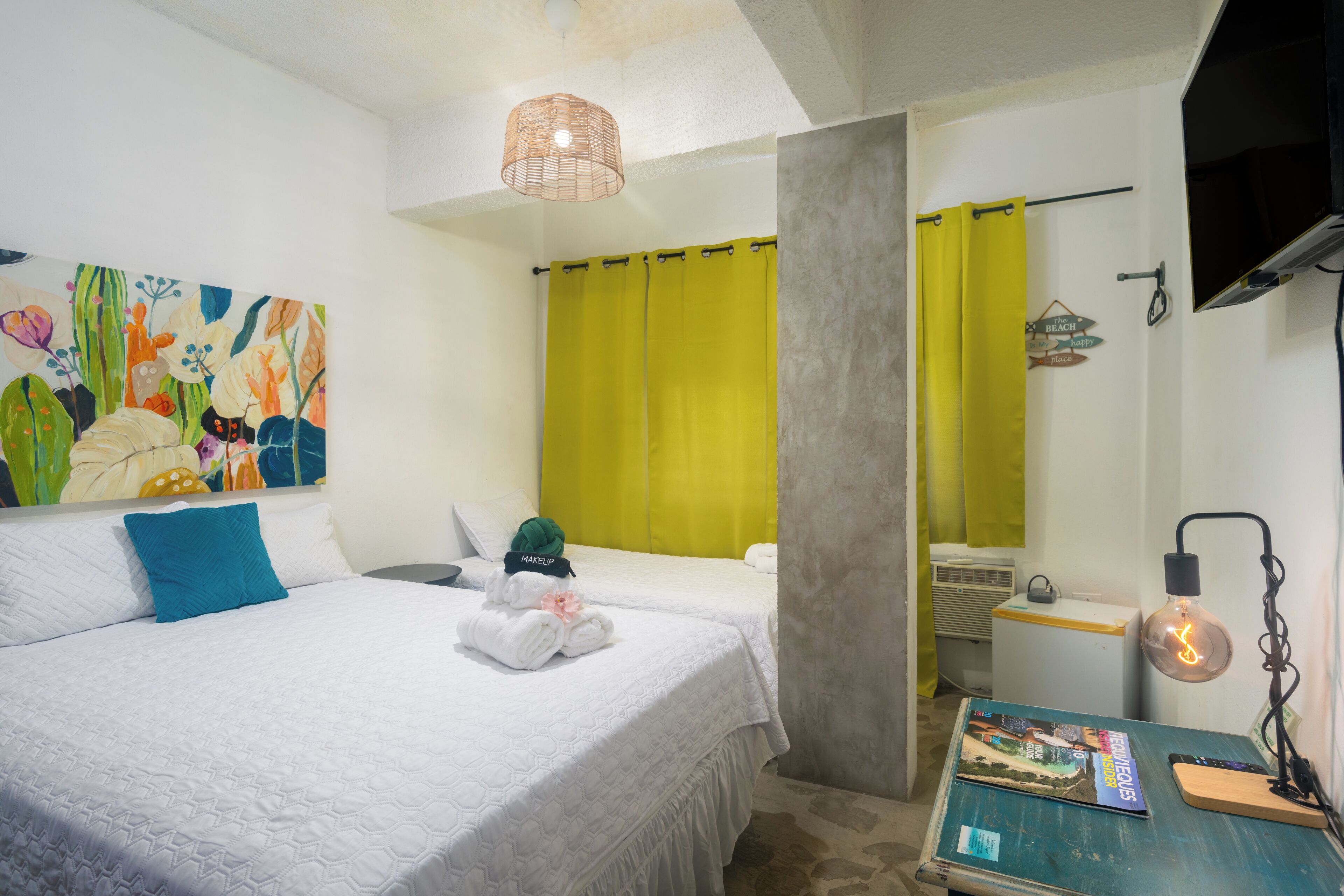 A hotel room with a bed, a desk with a magazine, a lamp, and a painting on the wall.