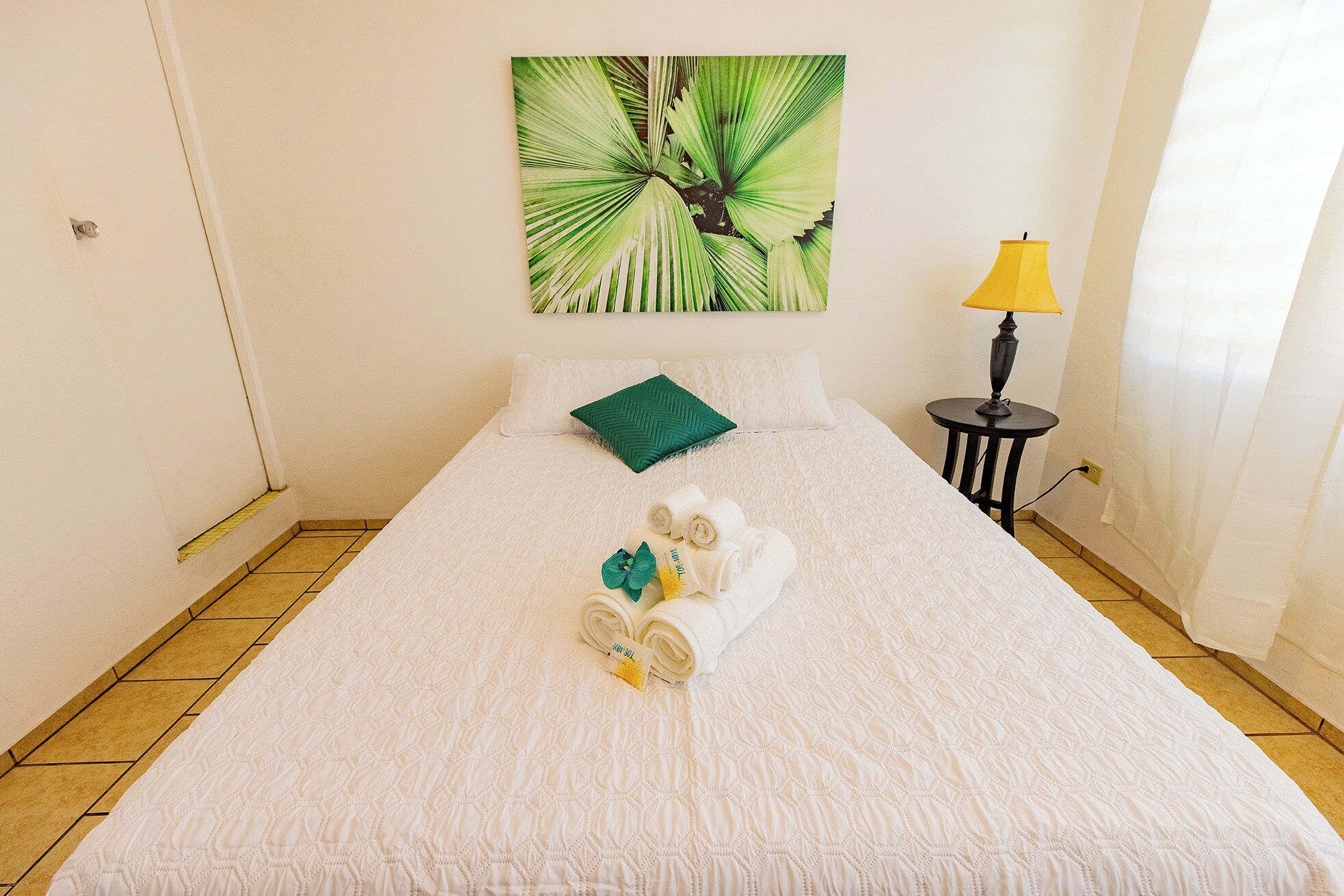 A neatly made bed with a white quilt, a green pillow, and rolled towels with a towel bar.
