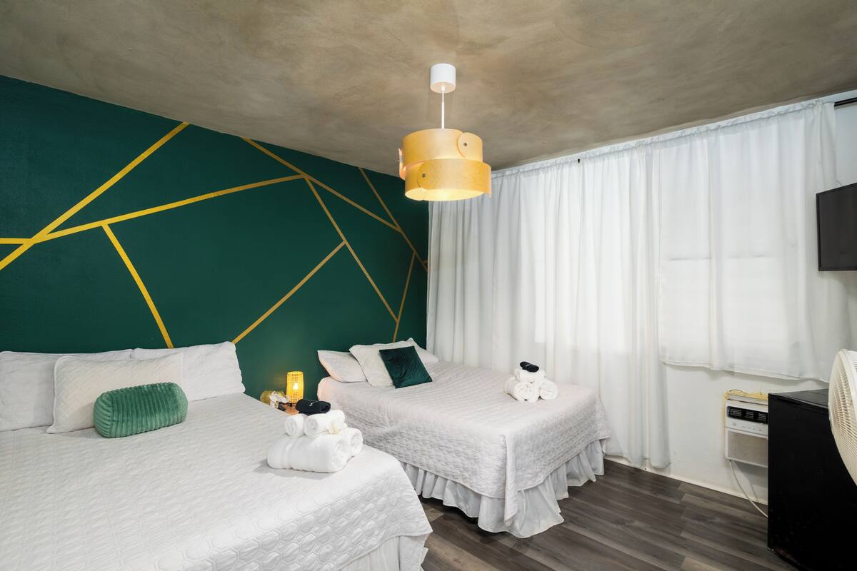 Ein Hotelzimmer mit zwei Betten, einem Wanddesign mit geometrischen Mustern in Grün, einer Deckenleuchte und einem Fernseher auf der rechten Seite.