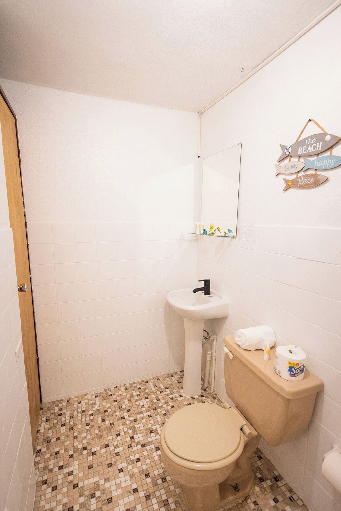 A small bathroom with a toilet, sink, and a mirror. There is a sign on the wall that says "The Beach Is My Happy Place."