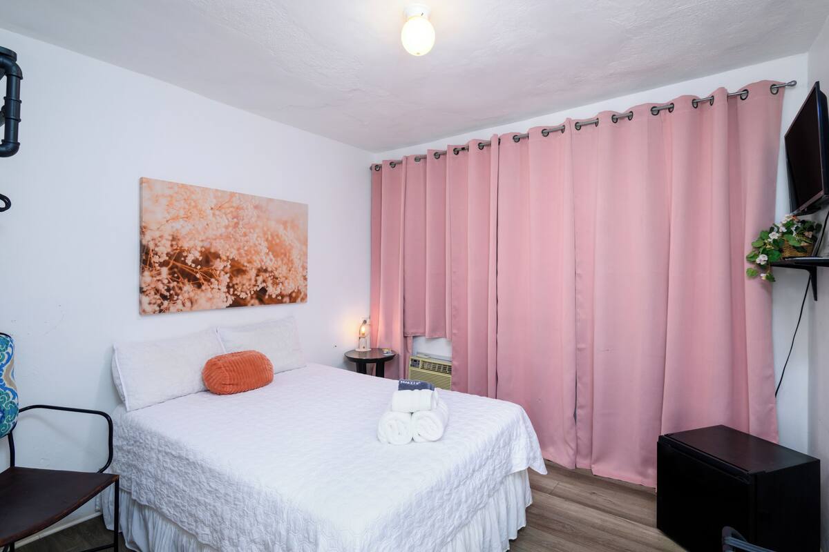 Ein ordentlich bezogenes Bett mit weißer Bettwäsche, einem rosa Vorhang und einem an der Wand hängenden Kunstwerk.