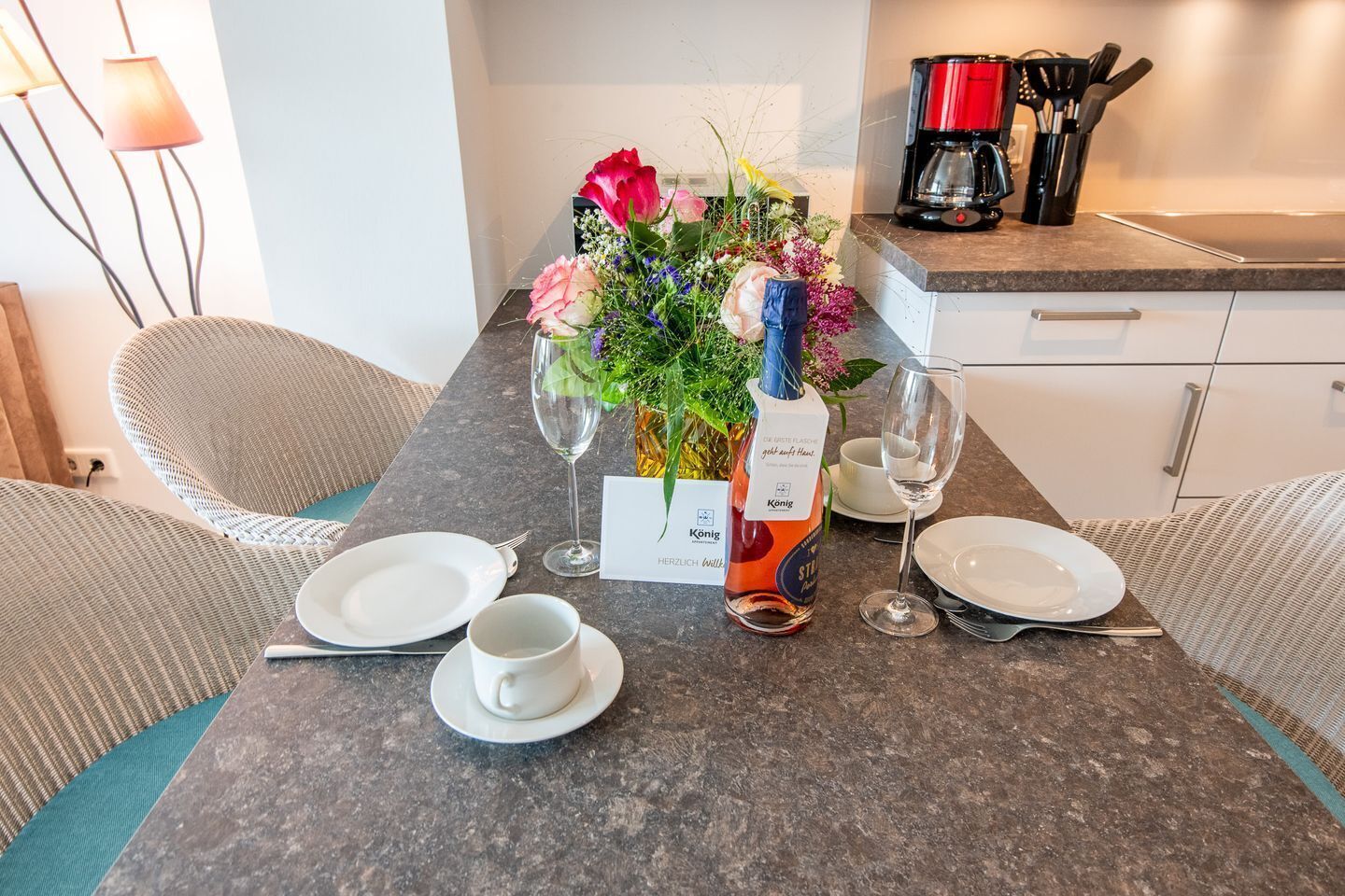 Una mesa de comedor dispuesta con platos, tazas y una botella de vino Konig, junto con un centro de mesa floral y una cafetera.