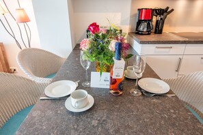 A dining table set with plates, cups, and a bottle of Konig wine, accompanied by a floral centerpiece and a coffee maker. - Haus Dünenweg Whg. 2 - Stilvoll. Modern. Weitblick. - Objekt-id: Odh/22 (Hörnum (Sylt))