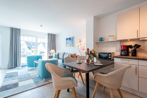 A modern living room with a sofa, chairs, and a dining table set for two. There is a kitchen area with a microwave and coffee maker.