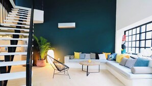 A modern living room with a white sofa, a round coffee table, and a black wireframe chair. There is a large window on the right, a potted plant, and a staircase on the left. - Casa Búho & Casa Arco (Tulum)
