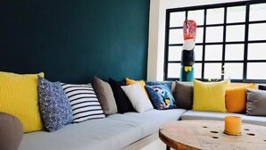 A modern living room with a grey sofa, colorful cushions, a wooden coffee table, and a large window with a view. - Casa Búho & Casa Arco (Tulum)