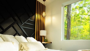 A bedroom with a bed, a nightstand, a lamp, a window with a view of trees, and a wall with a geometric pattern. - Rekindled @ The Patch, lux couples getaway, hot tub, outdoor living. (Laurelville)
