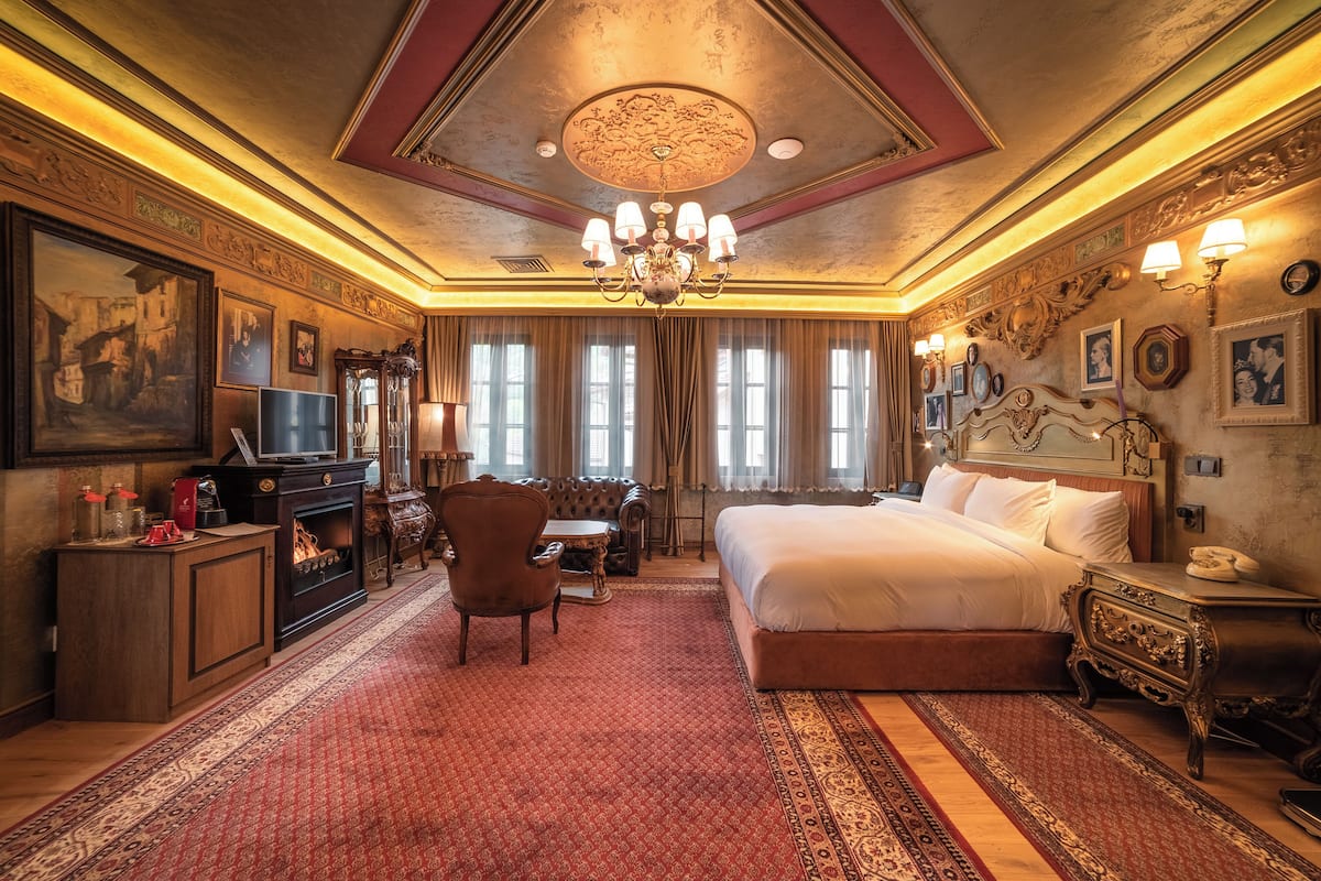 Ein Hotelzimmer mit einem großen Bett, einem Schreibtisch mit Lampe, einem Sessel, einem Kamin und einer Chandelier.