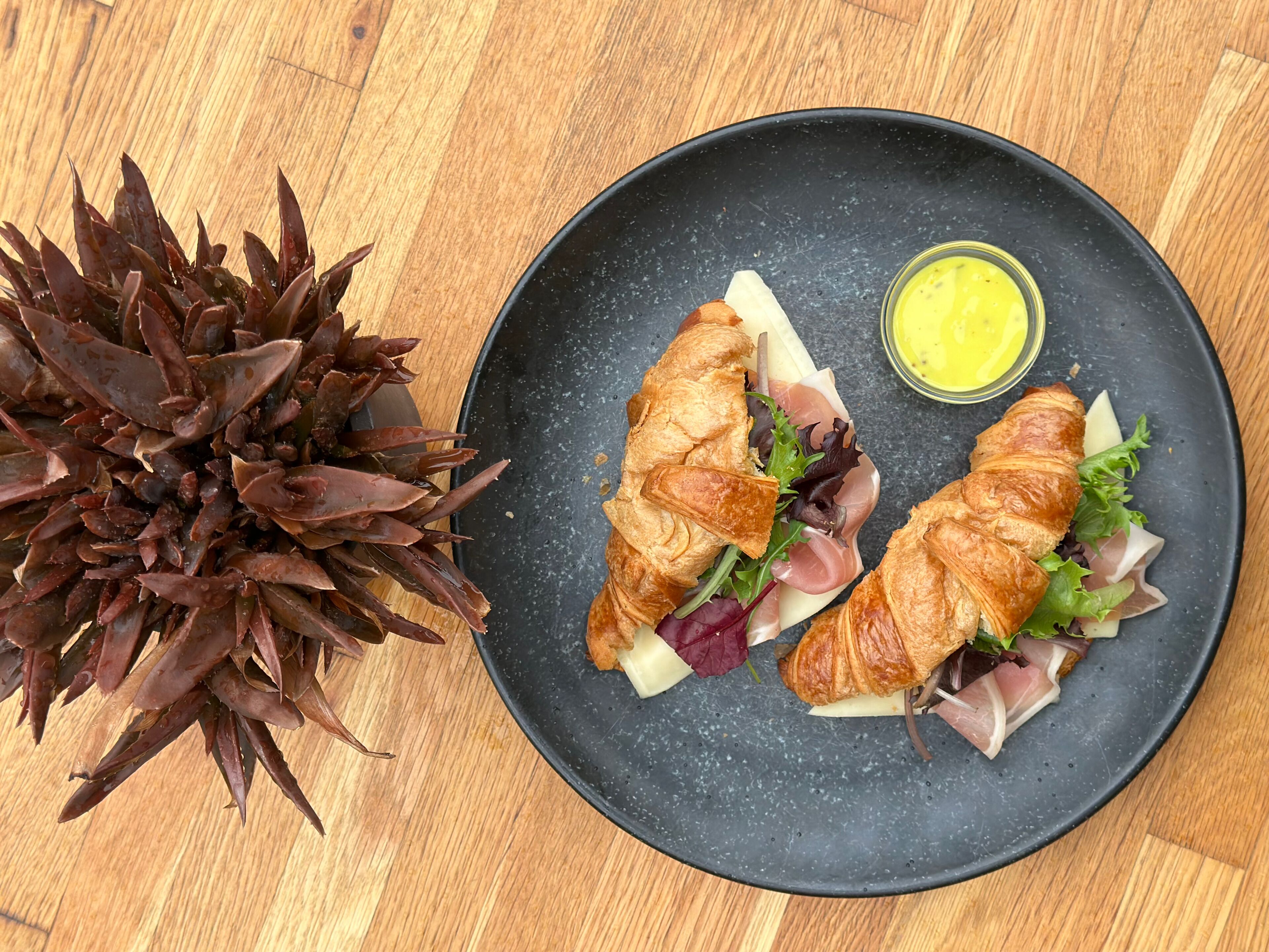A black plate with two croissants, a salad, and a small container of yellow sauce on a wooden table.