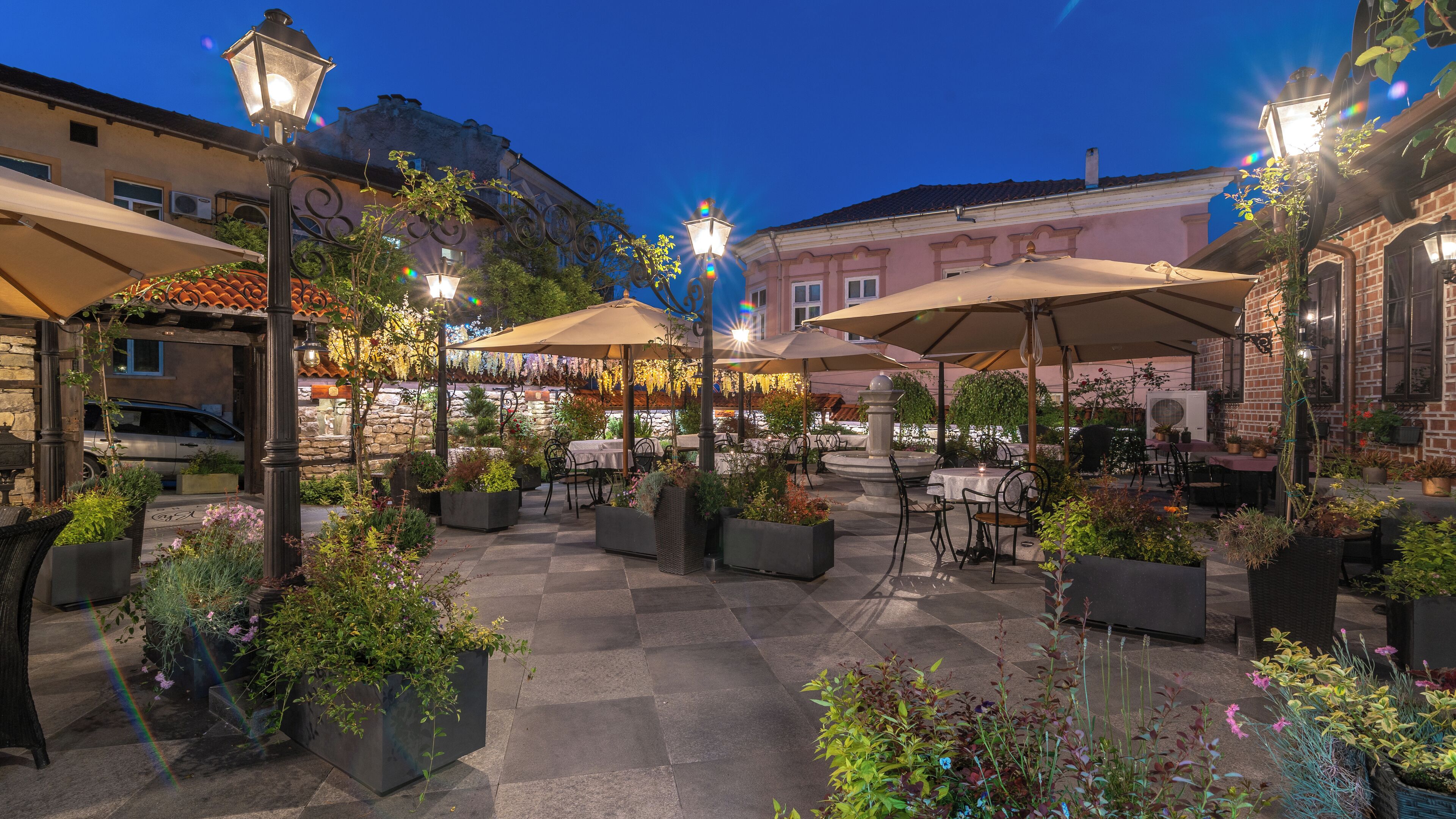 An outdoor patio area with tables and chairs, surrounded by potted plants and umbrellas.