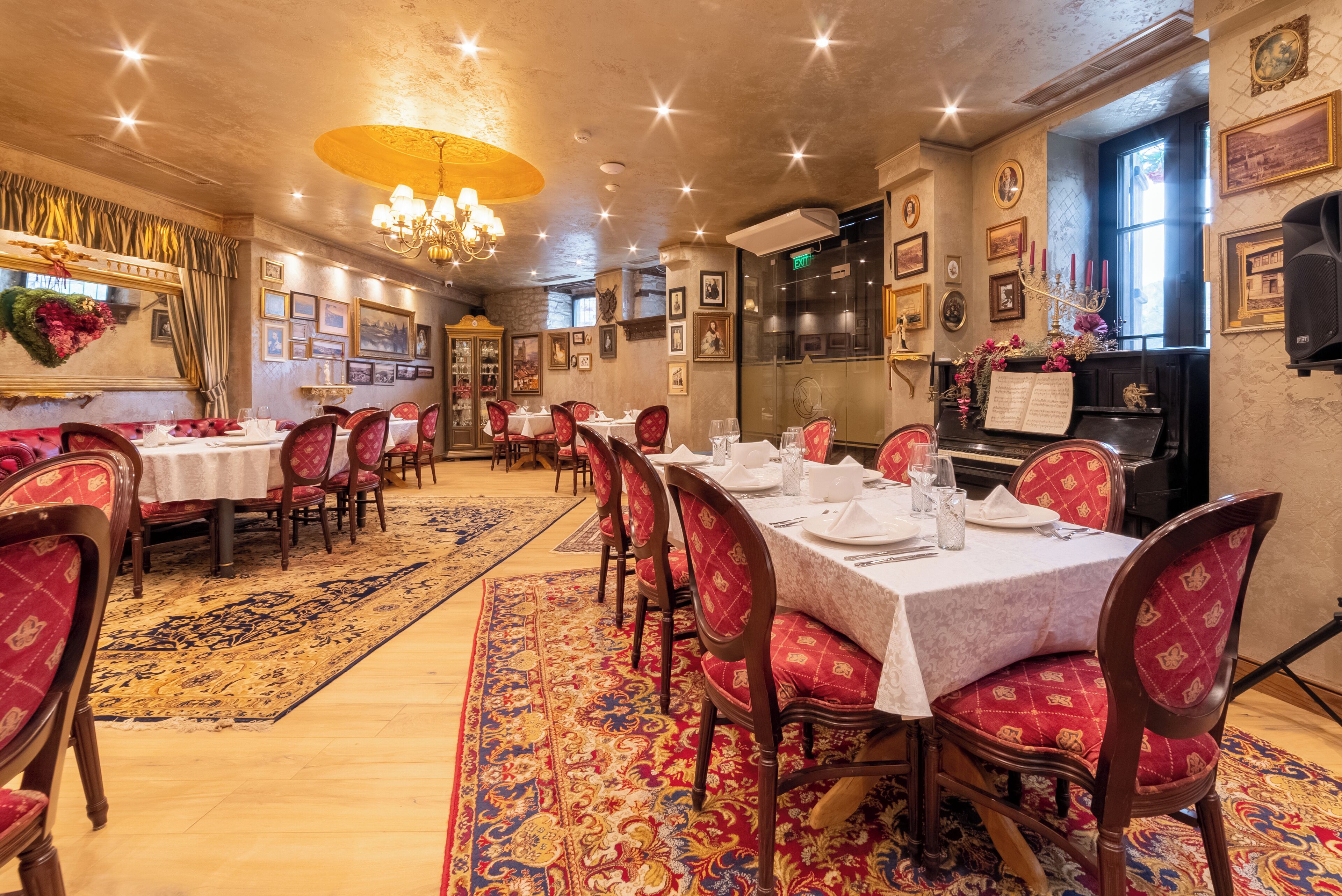 A dining area with tables set for service, red upholstered chairs, a chandelier, and a patterned carpet.