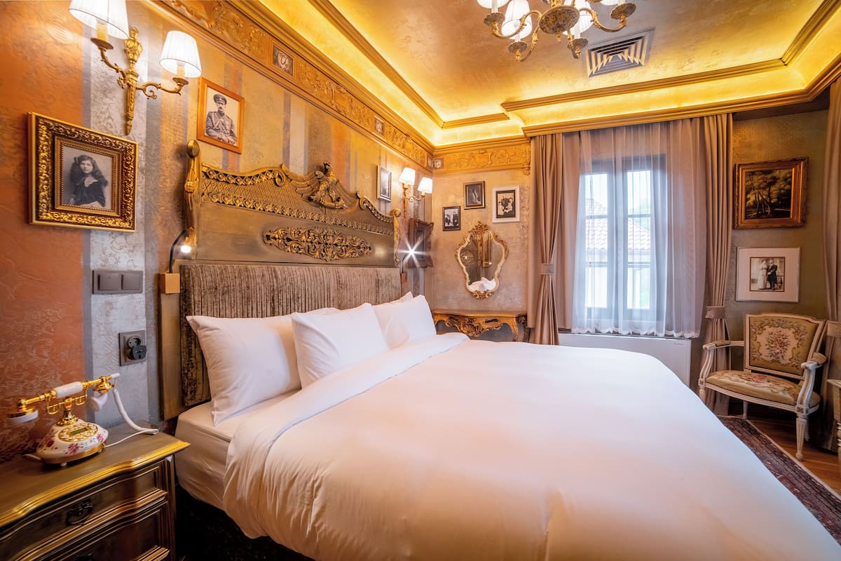 Ein luxuriöses Hotelzimmer mit einem großen Bett, einem aufwendig gestalteten Kopfteil und klassischer Einrichtung, einschließlich gerahmter Bilder und einem Vintage-Telefon.