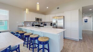 A modern kitchen with white cabinetry, a stainless steel refrigerator, a microwave, and a large island with bar stools. - Private Heated Pool, Pet Friendly, Parking for 5 (Port Aransas)