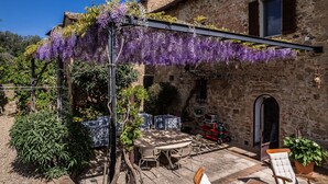 Un bâtiment rustique en pierre, agrémenté d’une pergola recouverte de fleurs violettes, d’un espace extérieur aménagé pour s’asseoir et d’un petit jardin.
