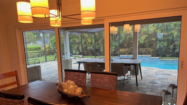 A dining area with a wooden table, wicker chairs, and a bowl of fruit. Outside, there is a pool and a patio with a table and chairs.