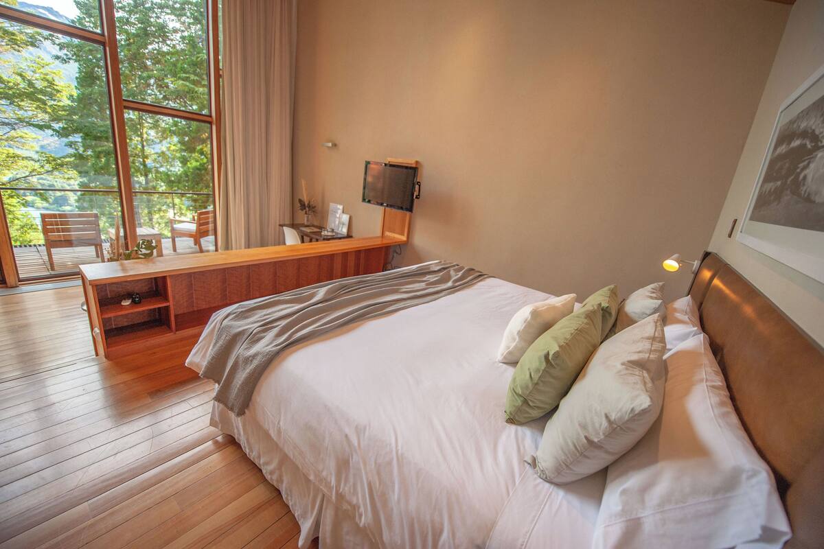 Un dormitorio con una cama grande, un escritorio de madera, una silla, un televisor y un balcón con vistas a árboles.