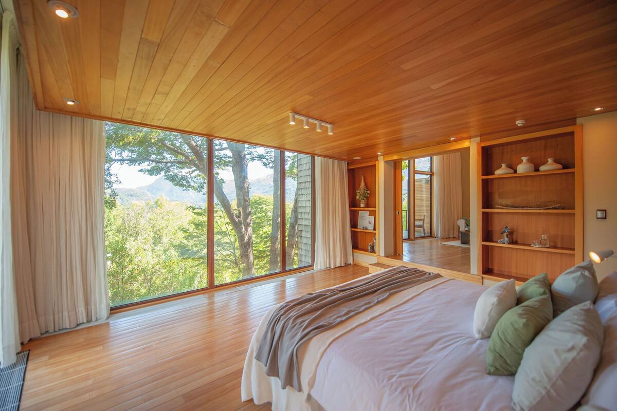 Un dormitorio amplio con una cama grande, suelo de madera, estantería empotrada y un ventanal con vistas a árboles y montañas.