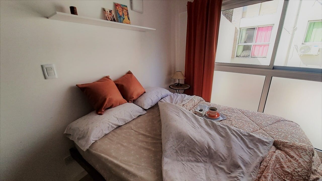Ein Bett mit gemustertem Überwurf, zwei Kissen, ein kleiner Tisch mit Lampe und ein Fenster mit Vorhängen.