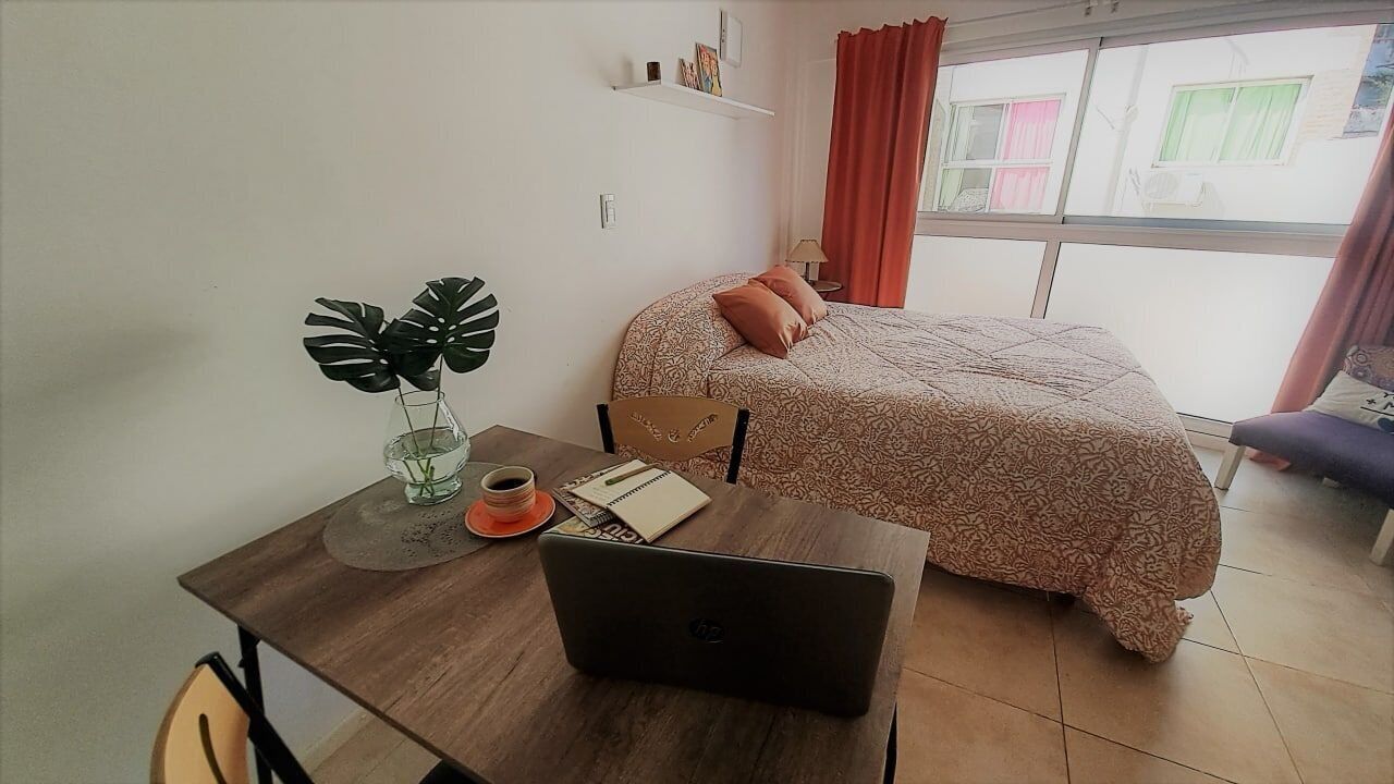 Quarto com cama, um laptop sobre mesa de madeira, uma planta em vaso e uma janela com cortinas.