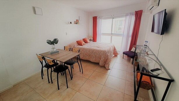 Una habitación pequeña y luminosa con una cama, un escritorio con sillas, un televisor y una ventana con cortinas.