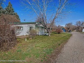 A single-story house with a green roof and a front porch, surrounded by trees and a gravel driveway. - An Oasis inside and out! Sweet cottage in Silt (Silt)
