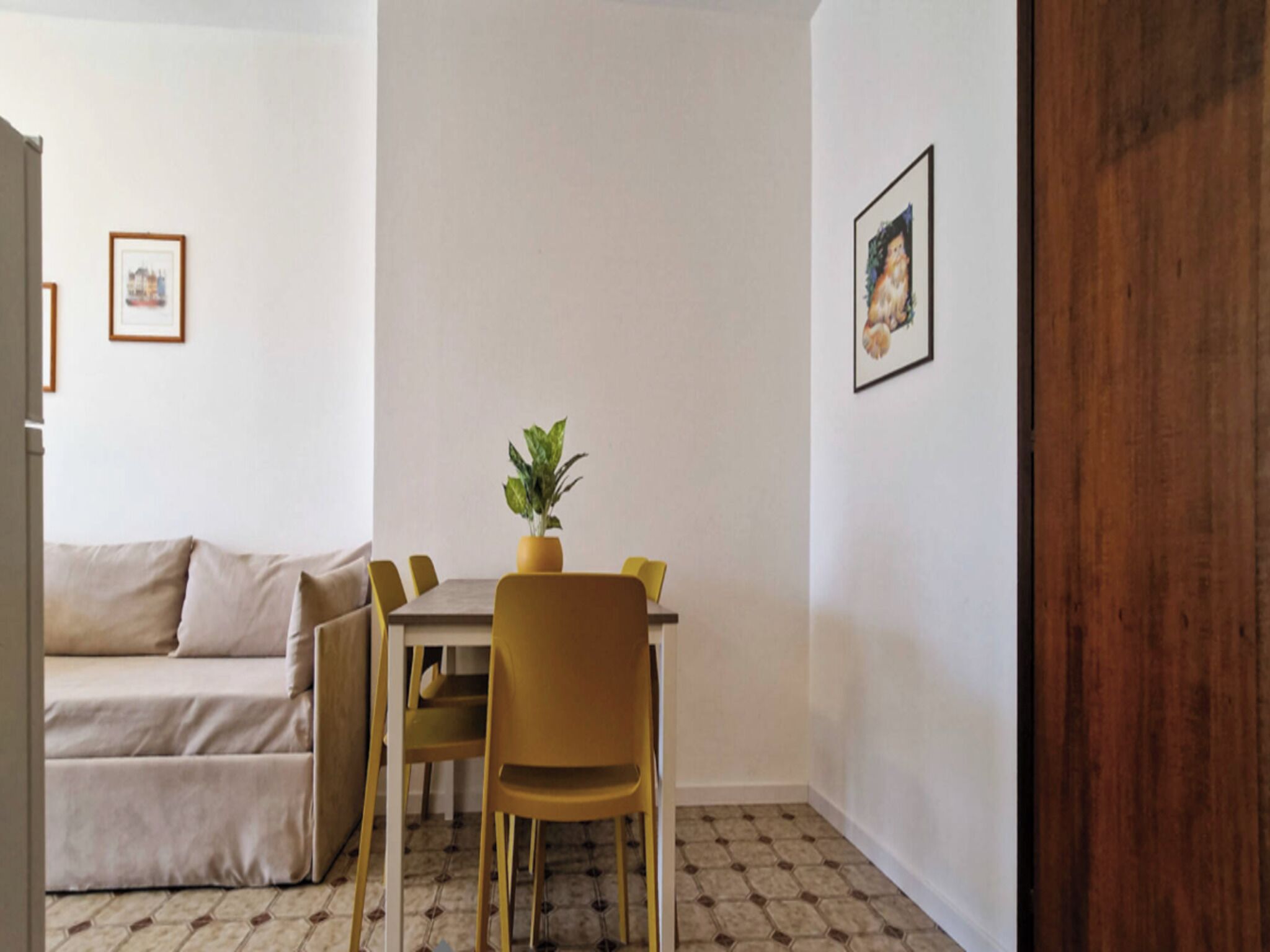 A small, neatly arranged room with a sofa, a dining table with chairs, a potted plant, and framed pictures on the walls.