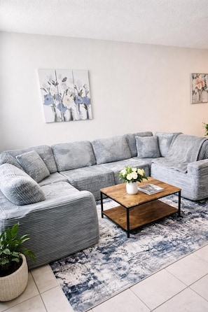 A grey sectional sofa, a wooden coffee table with a vase of flowers, a patterned rug, and a wall with a floral painting.