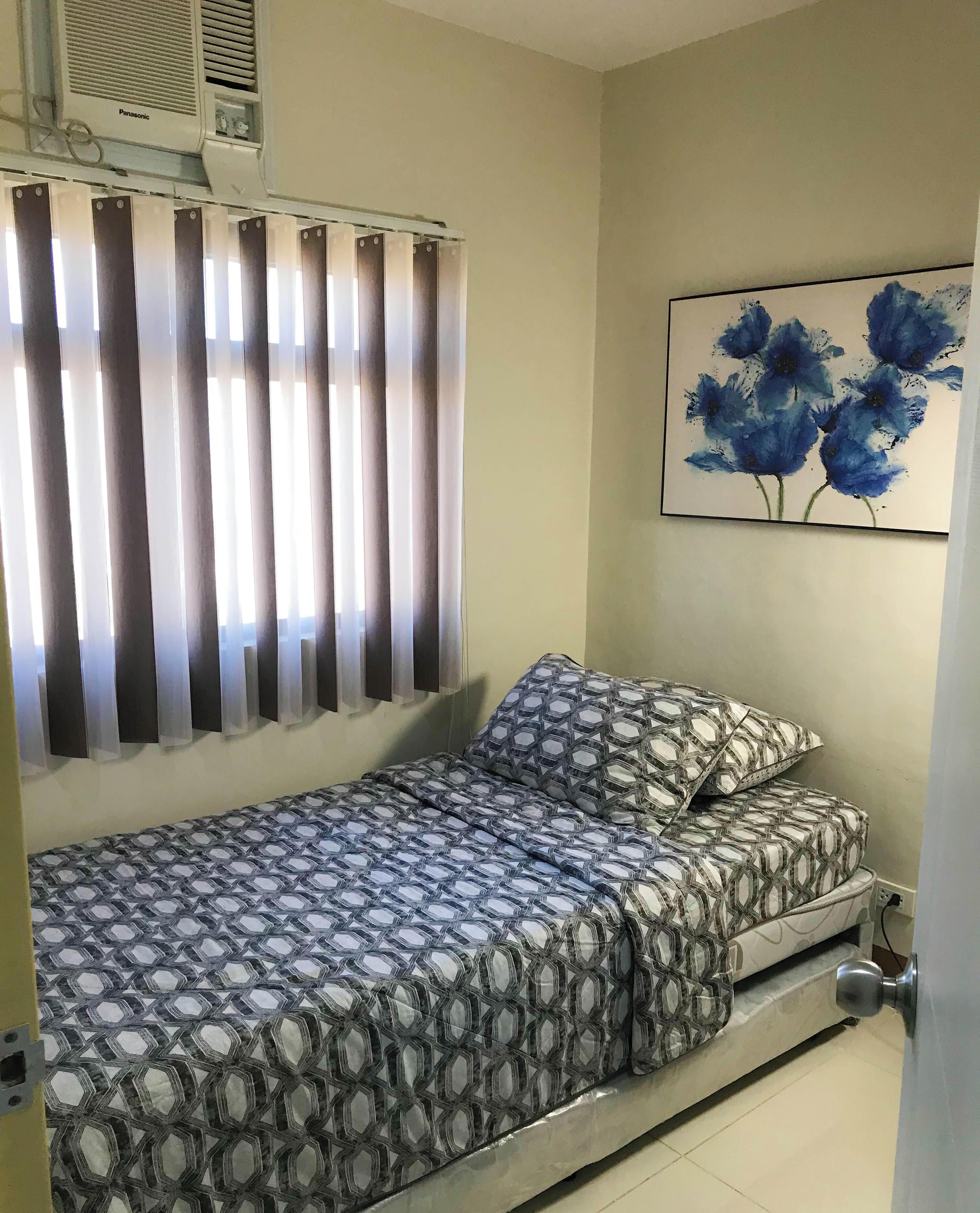 A single bed with patterned bedding, a wall-mounted air conditioner, a window with vertical blinds, and a painting of blue flowers on the wall.
