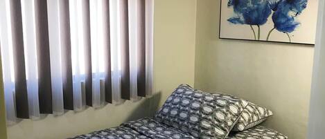 A single bed with patterned bedding, a wall-mounted air conditioner, a window with vertical blinds, and a painting of blue flowers on the wall.
