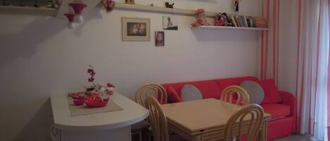 Un coin repas avec une table et des chaises, un canapé rouge et des étagères ornées d’objets décoratifs.