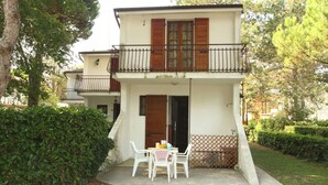 A two-story building with a balcony, a table with chairs, and a hedge in the foreground. - Welcoming Accommodation in Bibione (San Michele al Tagliamento)