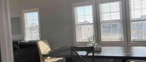 Ein modernes Wohnzimmer mit einem dunklen Sofa, einem Esstisch mit Stühlen und großen Fenstern mit Jalousien.