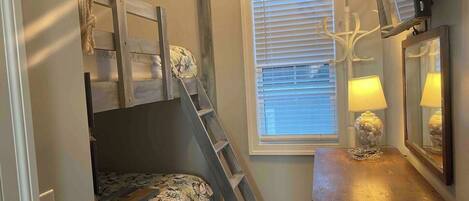 A bedroom with a bunk bed, a ladder, a bedside table with a lamp, and a window with blinds.