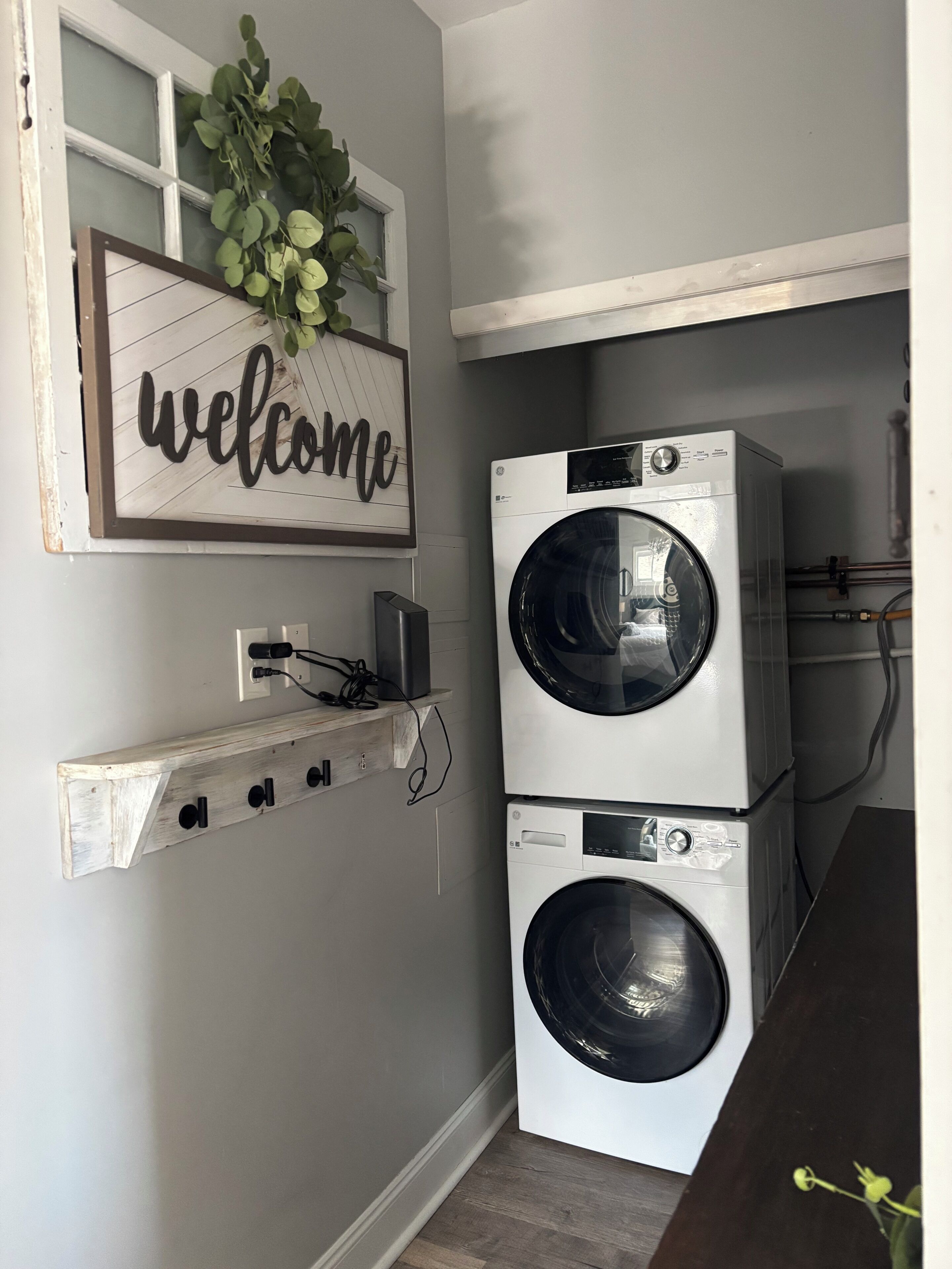A laundry room with two stacked front-loading washing machines, a 'welcome' sign, and a power outlet with a cable.
