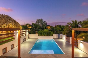A swimming pool with a wooden deck, surrounded by plants and a thatched roof structure.