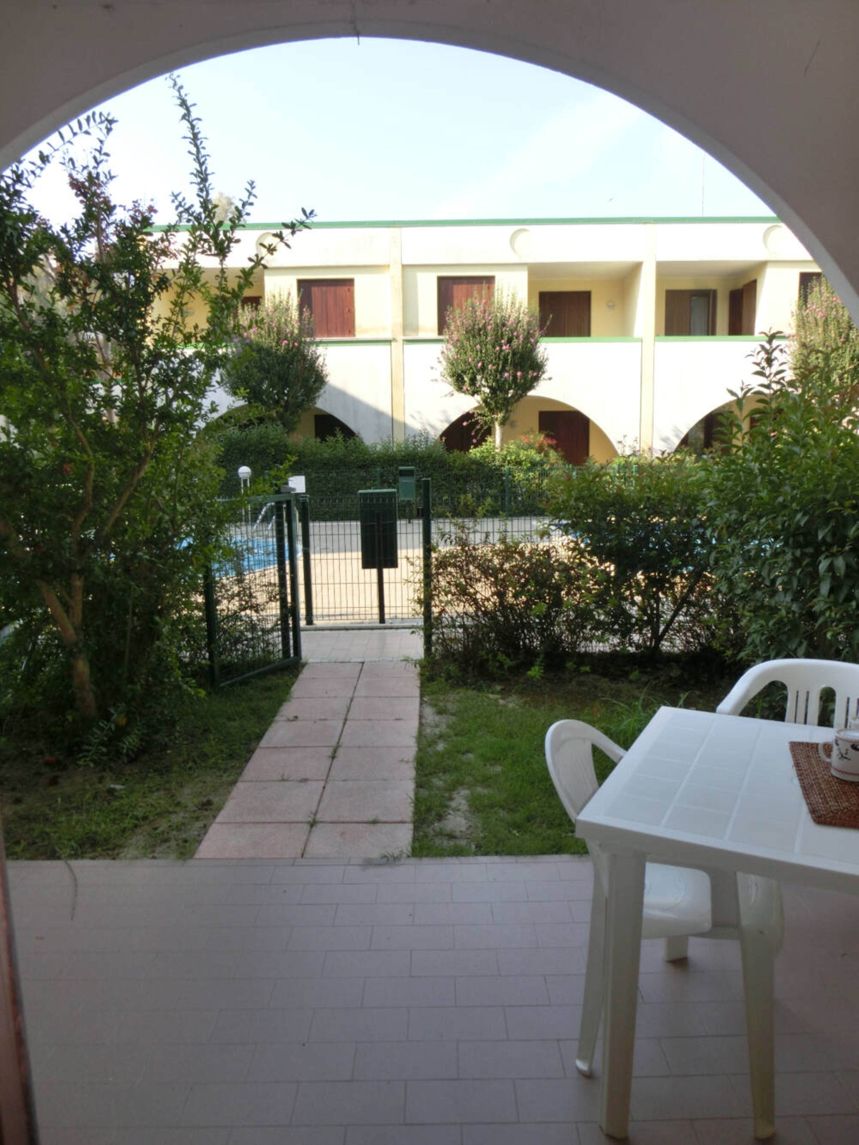 An outdoor patio area with a white table and chairs, a pathway leading to a building with a green roof, and a fenced garden area.