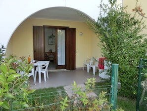 Un patio cubierto con una mesa y sillas, una puerta que da a una habitación interior, y una cerca verde.