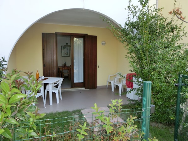 Une terrasse couverte avec une table et des chaises, une porte donnant sur une pièce intérieure et une clôture verte.